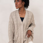 Model looking to the side while wearing the Chunky Knit Cable Cardigan in Almond paired with white pants