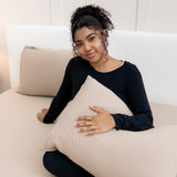 Model sitting on bed overtop the Fitted Sheet in Almond holding a pillow in an Almond Pillowcase