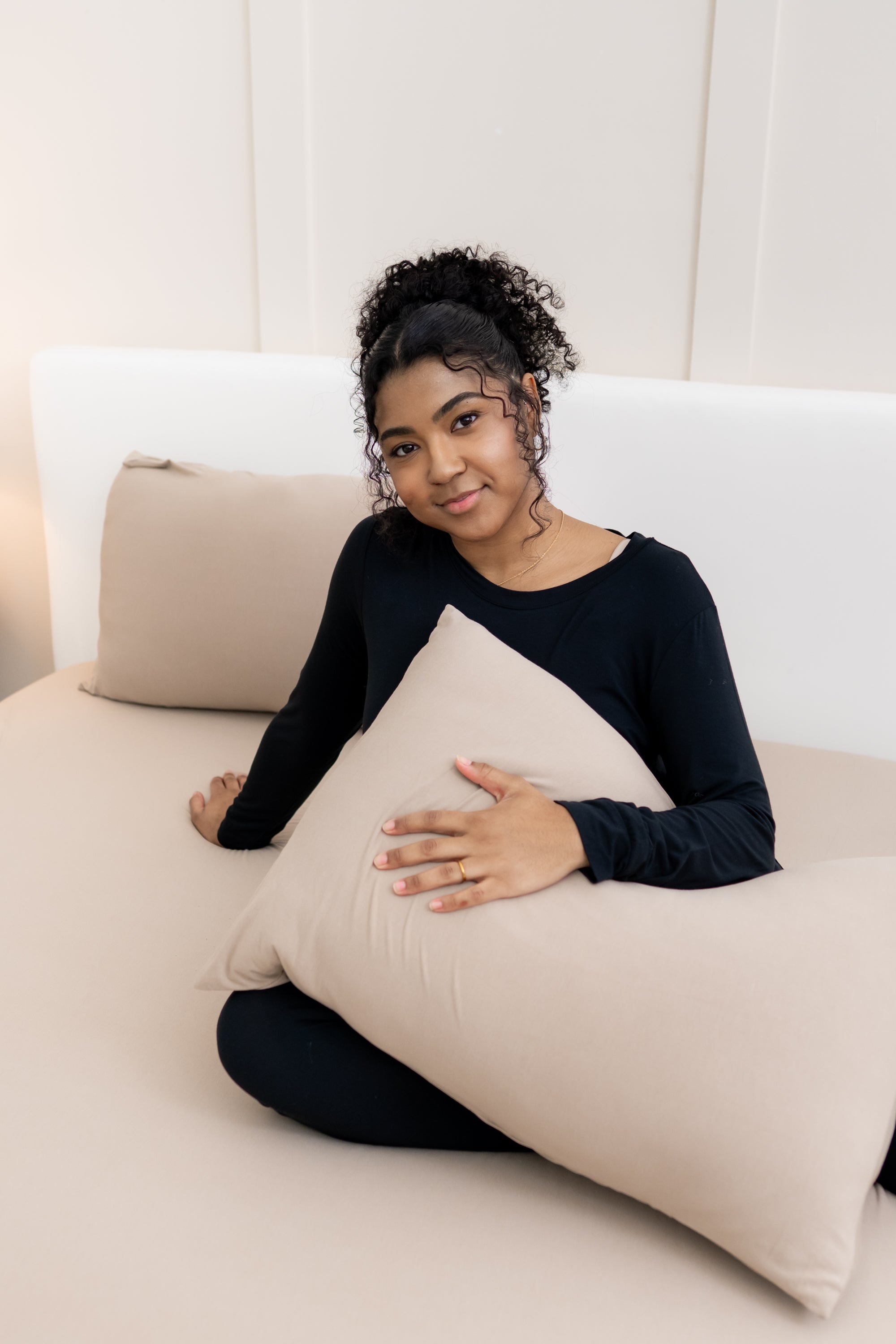 Model sitting on bed overtop the Fitted Sheet in Almond holding a pillow in an Almond Pillowcase