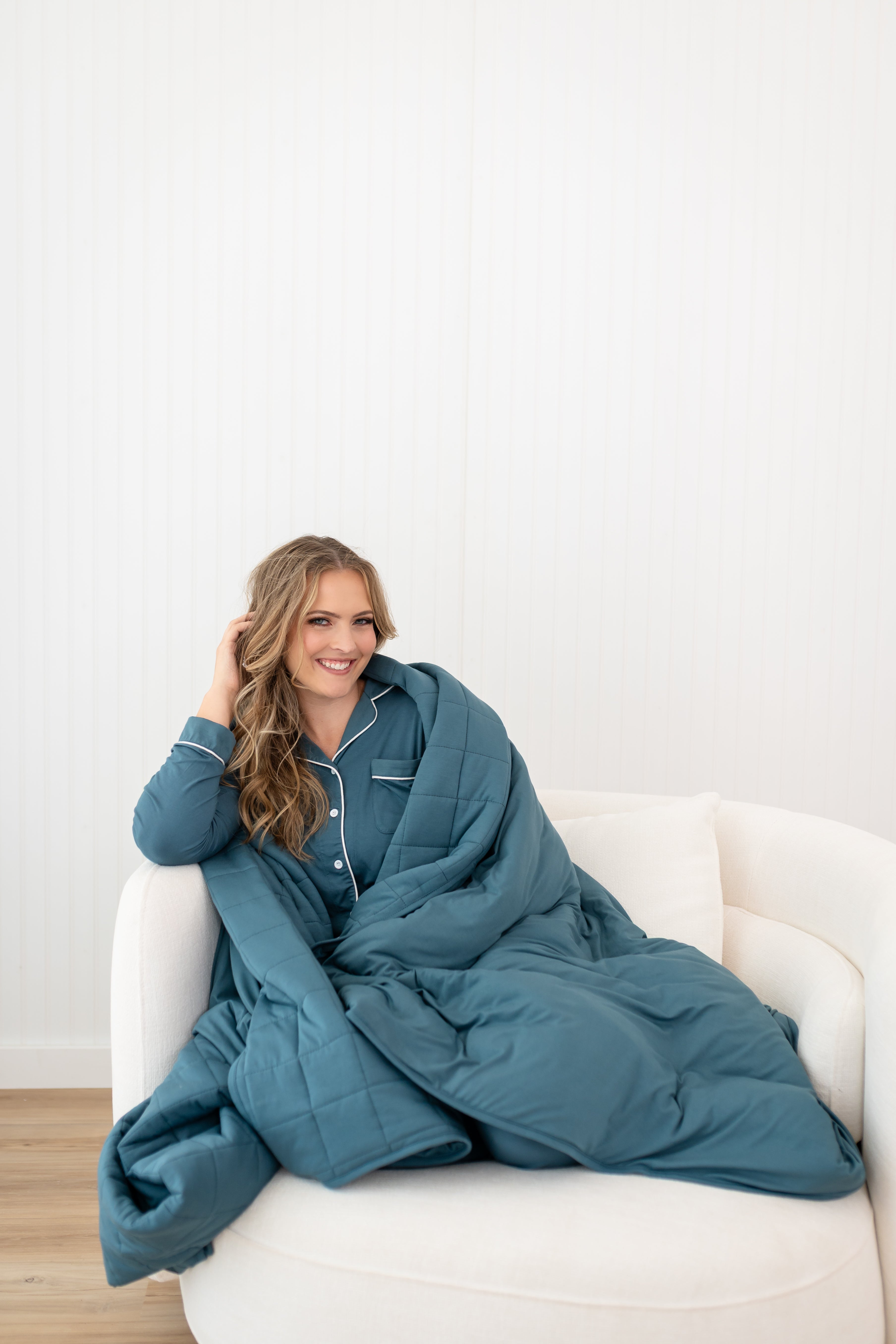 Smiling model sitting on a white chair while wrapped in the Quilt Blanket in Atlantic 2.5 TOG