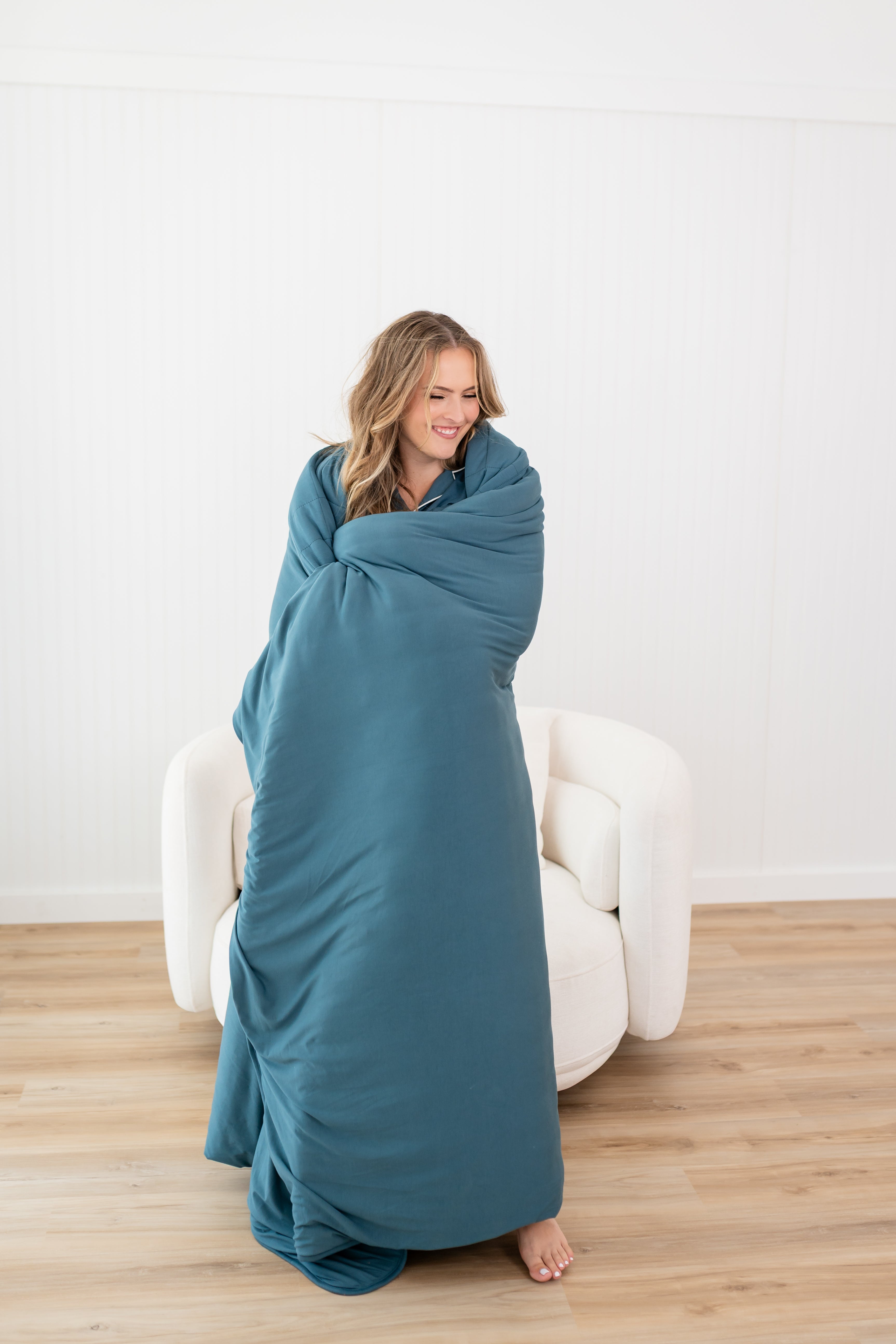 Standing and smiling model wrapped in the Kyte Quilt Blanket in Atlantic 2.5 TOG