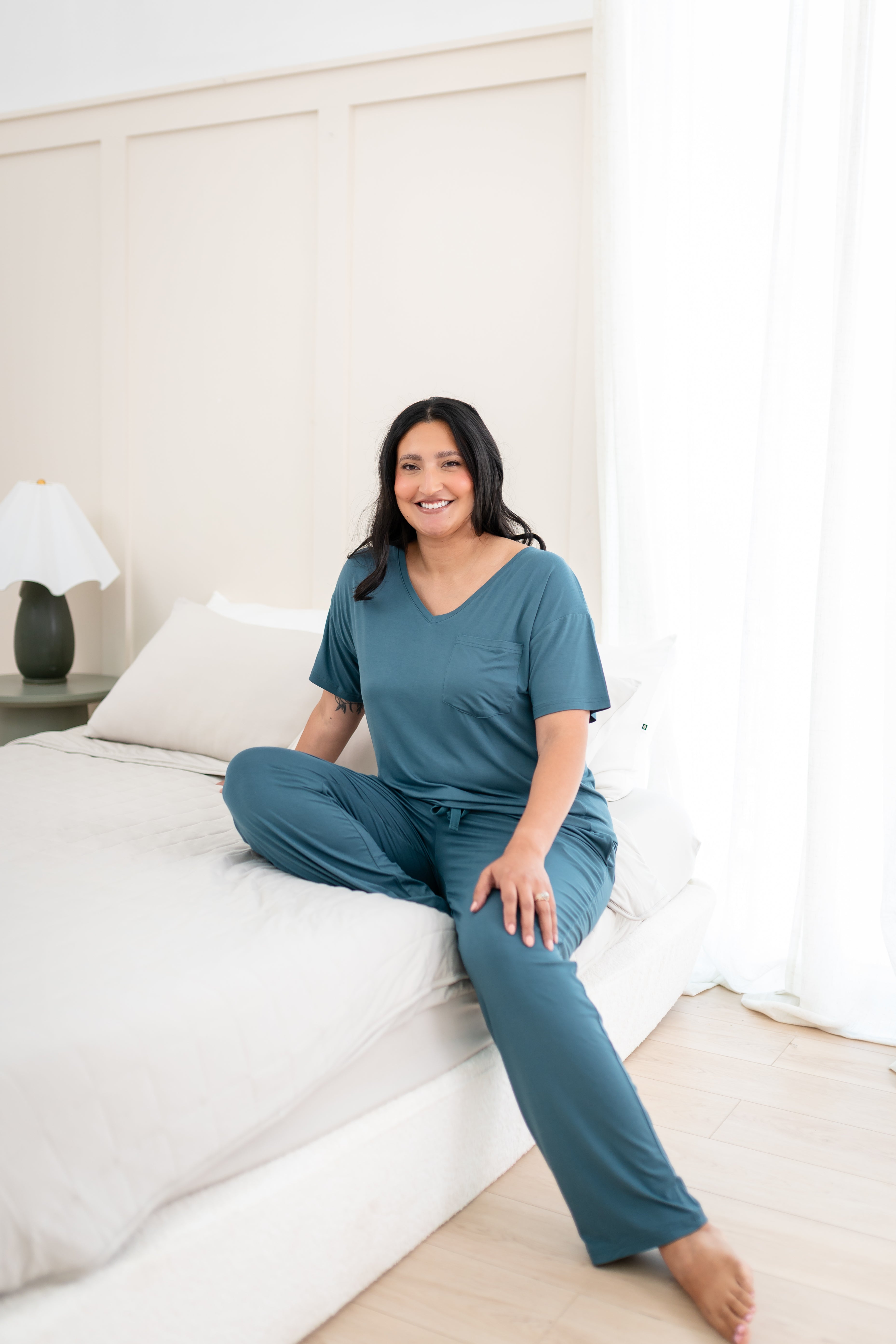 Smiling model sitting on side of bed while wearing Women's Lounge Pants in Atlantic