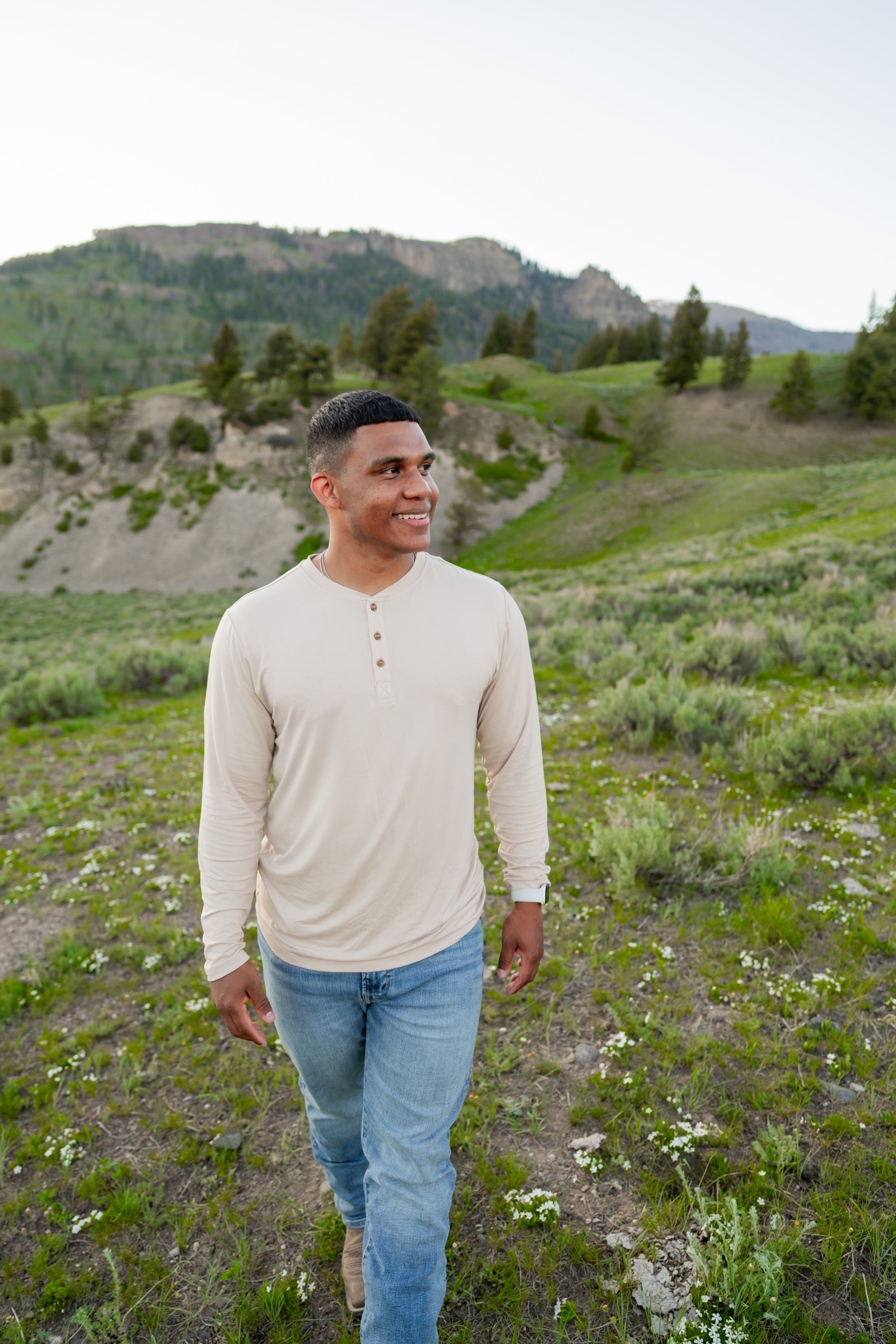Model walking in grassy field with mountains in background while wearing Men's Long Sleeve Henley in Bisque