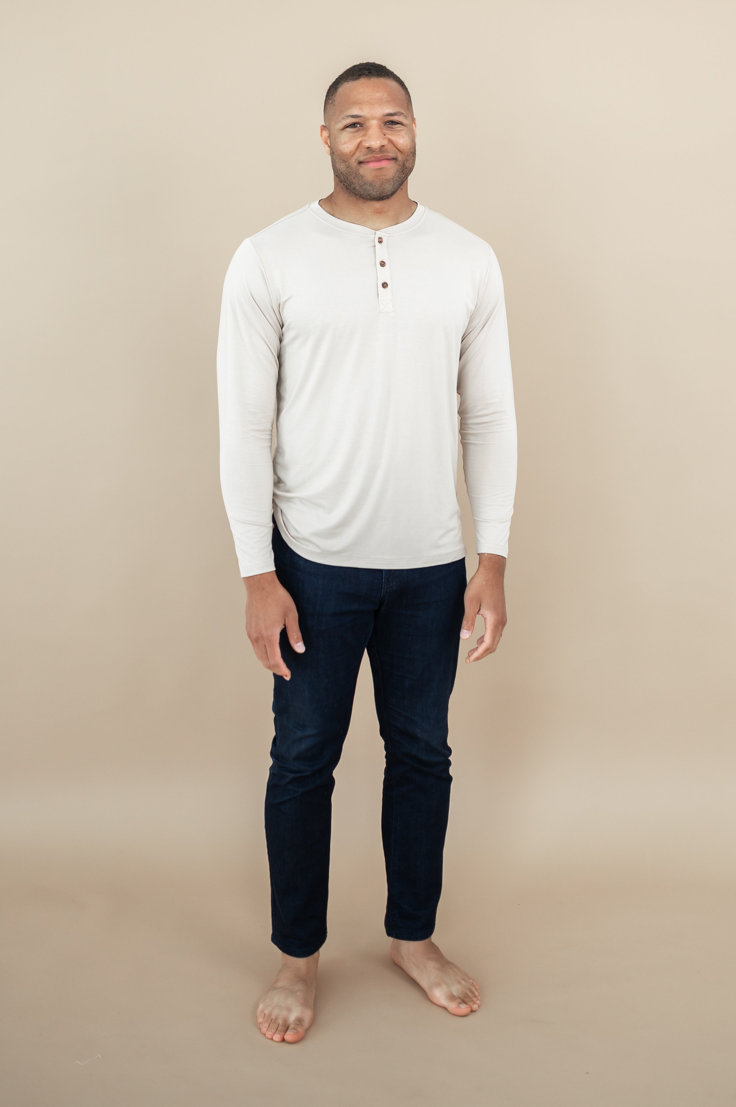Model standing in tan rom while wearing the Kyte Men's Long Sleeve Henley in Bisque paired with dark-wash jeans