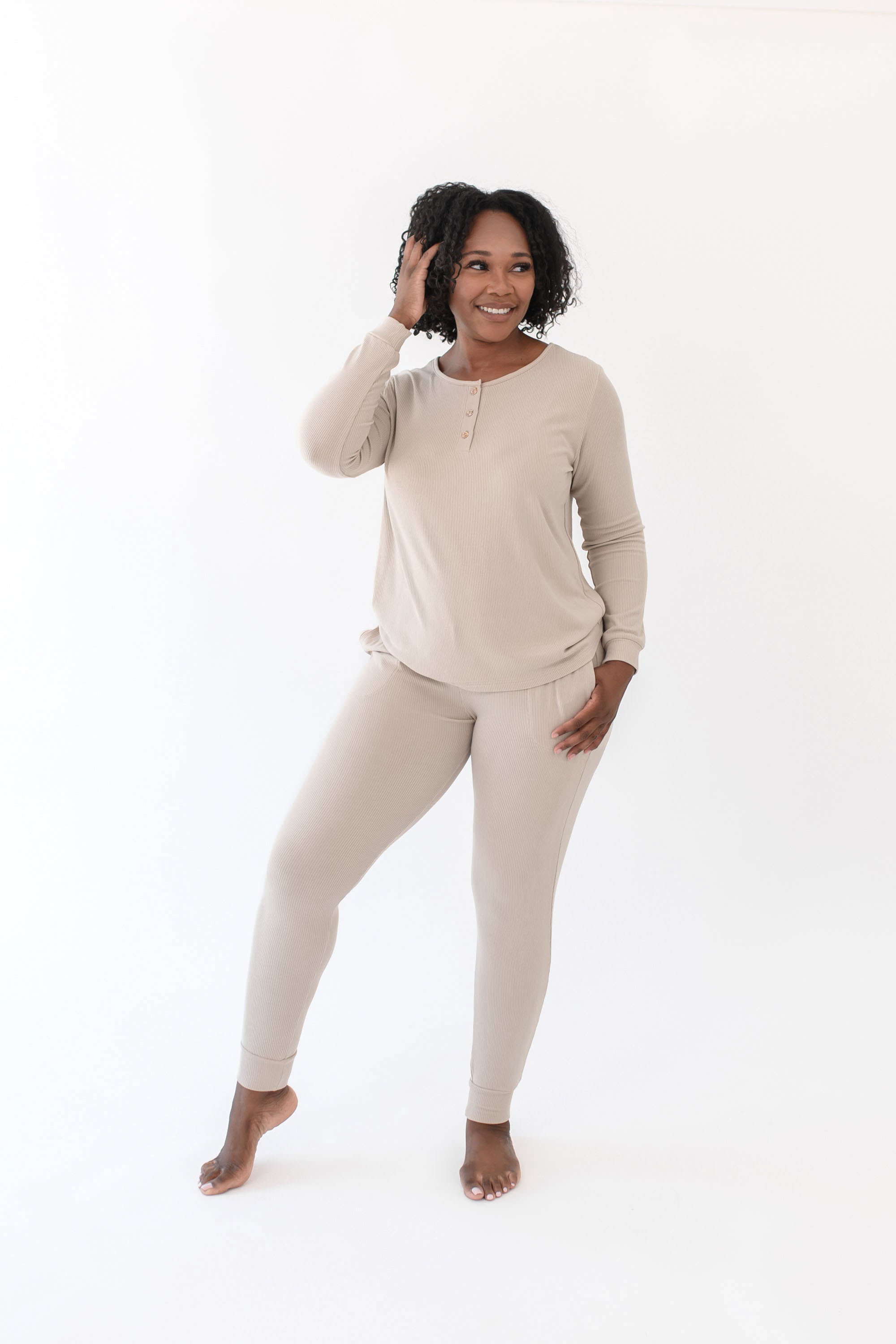 Smiling female standing with one hand in her hair and the other hand in the pants pocket of the Women’s Ribbed Henley Set in Bisque