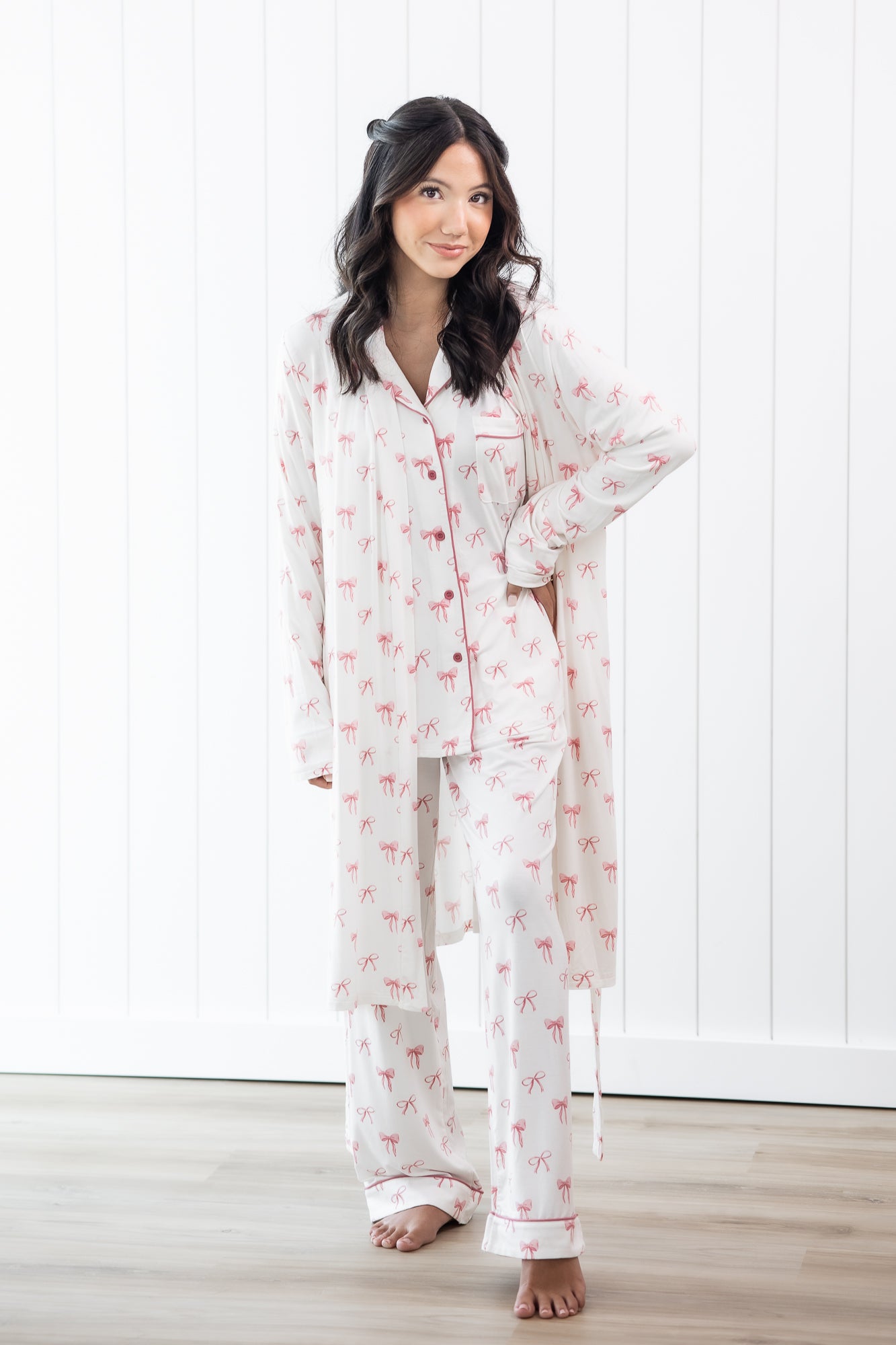 Woman wearing a white pajama set with pink bow patterns in a room with white walls and wooden floor.