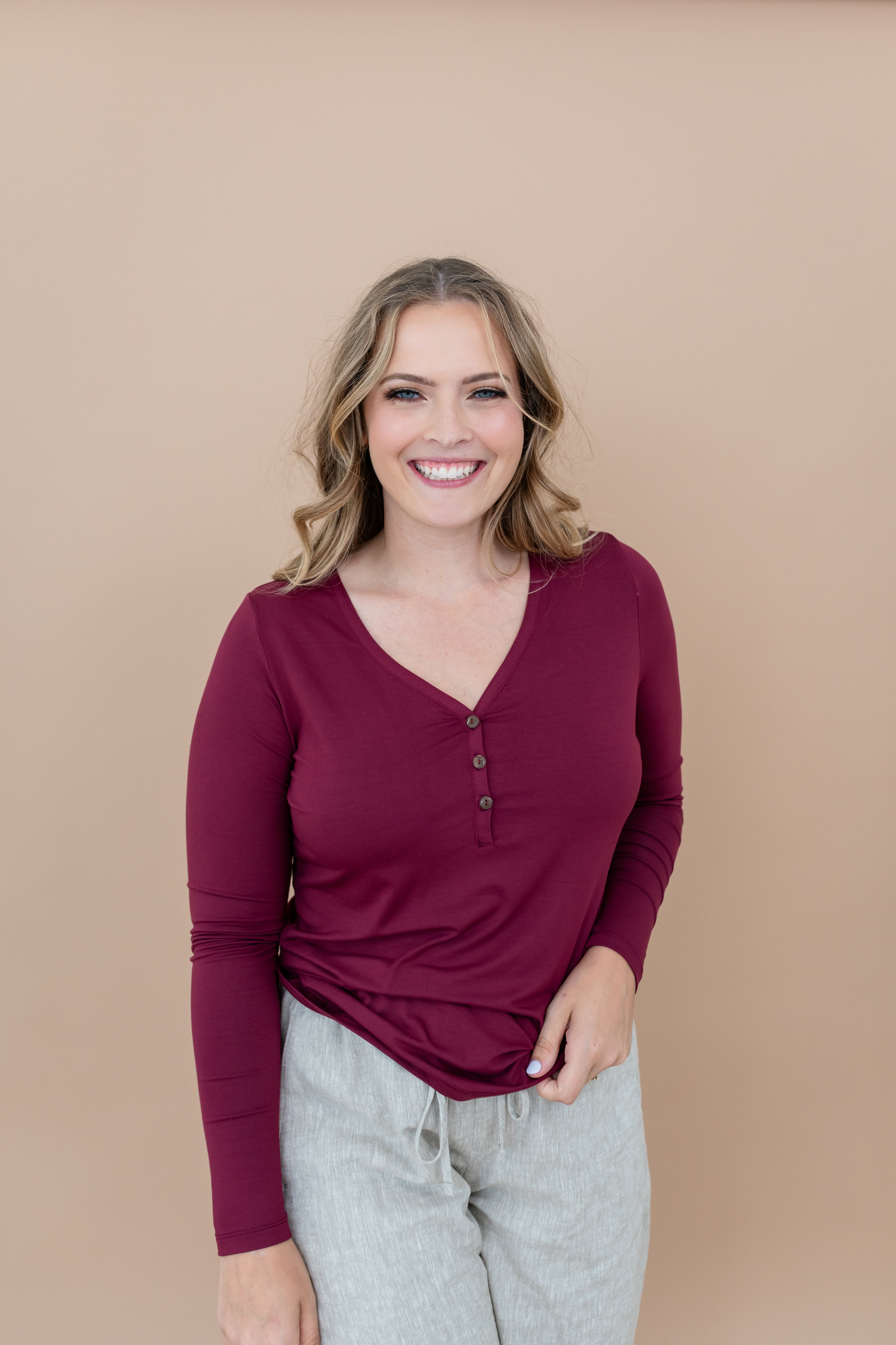 Woman smiling while modeling Henley top in Burgundy with hand on hip and paired with light bottoms