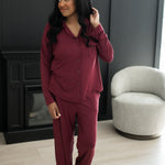 Woman standing in front of a black wall and fireplace while modeling the Bamboo Long-Sleeved Women's Pajama Set in Burgundy with Burgundy Trim