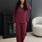 Woman standing in front of a black wall and gray chair while modeling the Kyte Bamboo Long-Sleeved Women's Pajama Set in Burgundy with Burgundy Trim