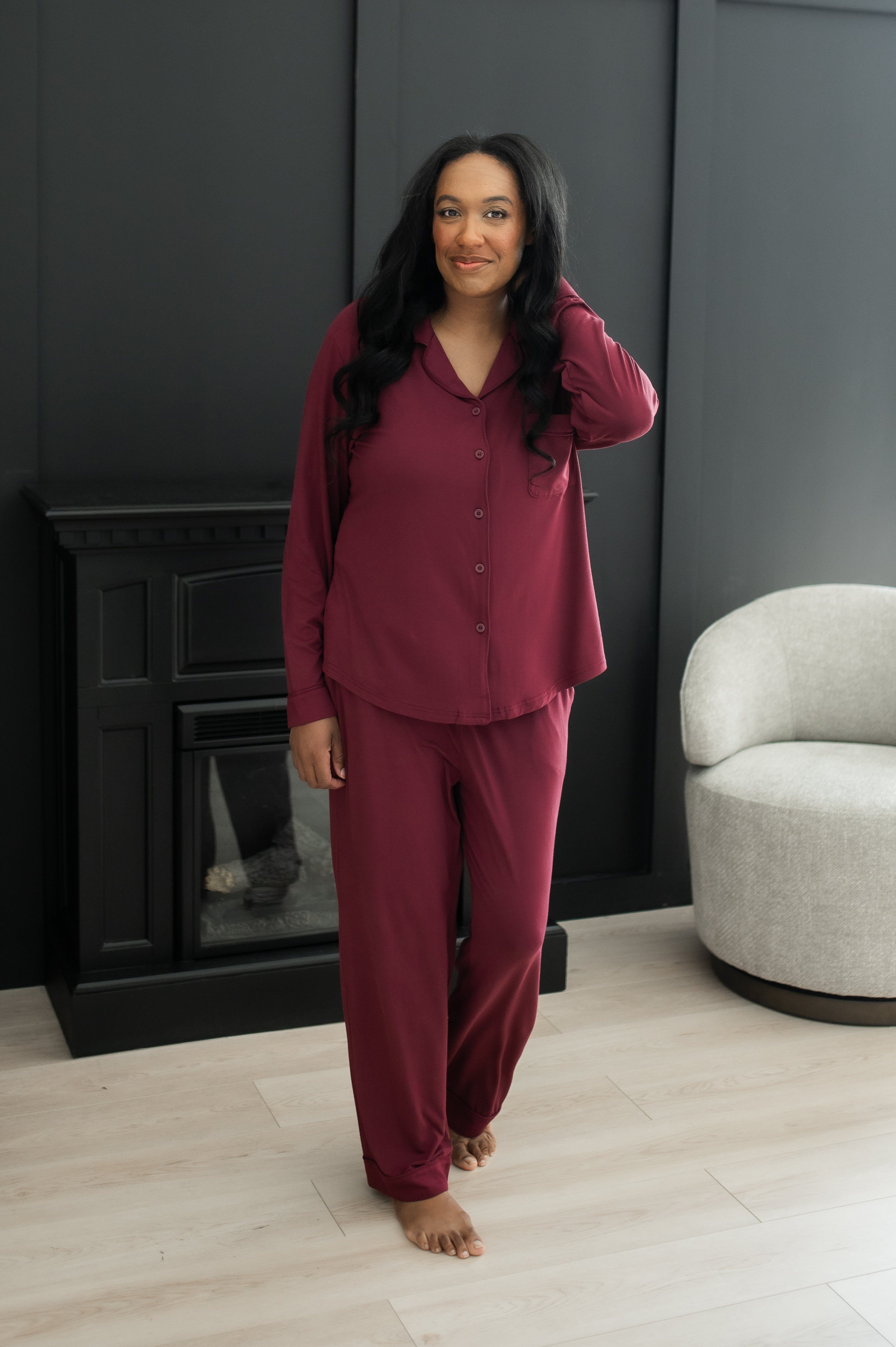 Woman standing in front of a black wall and gray chair while modeling the Kyte Bamboo Long-Sleeved Women's Pajama Set in Burgundy with Burgundy Trim