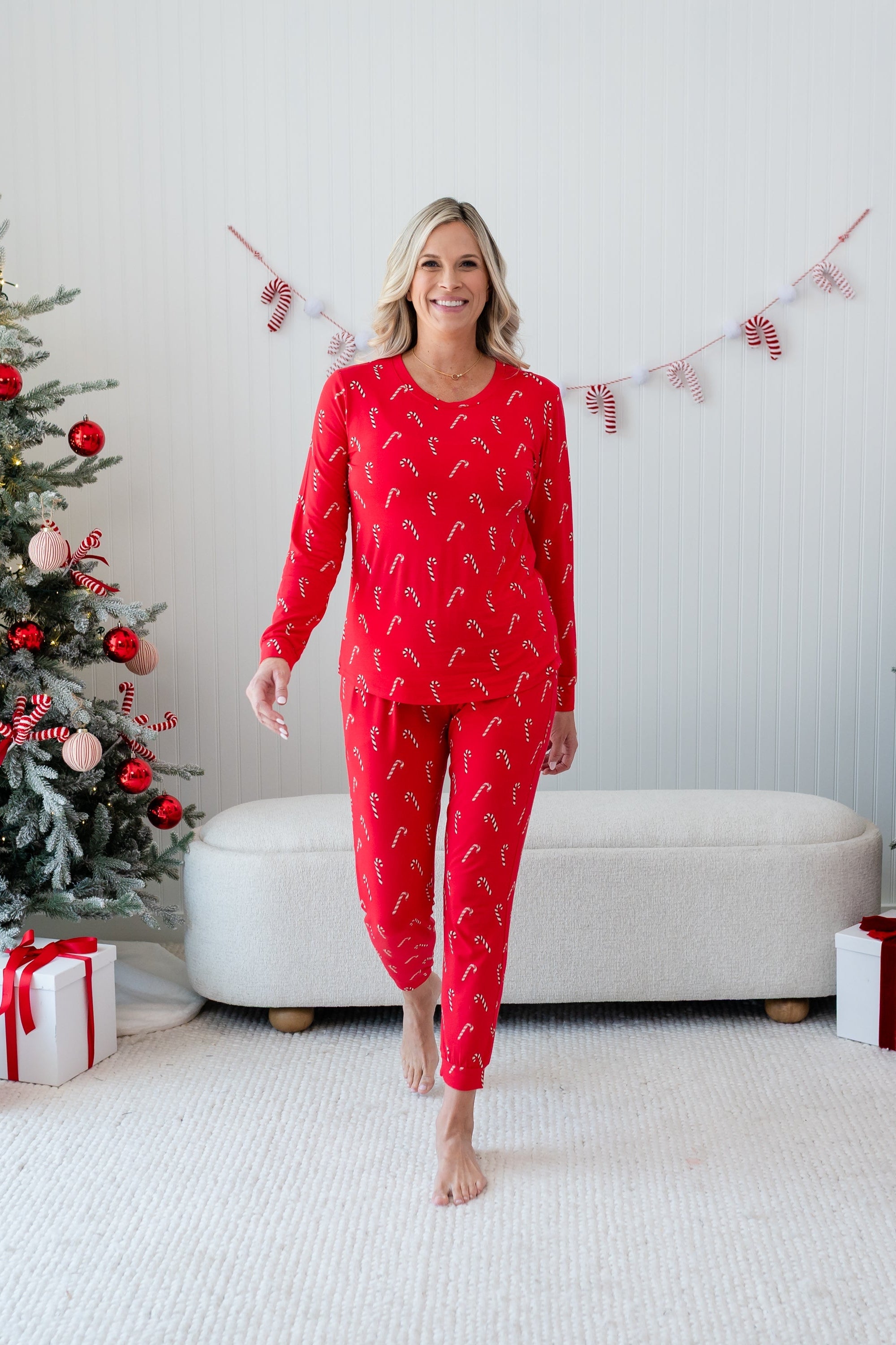 Smiling female model walking away from two decorated christmas trees and a beige ottoman wearing the Women's Jogger Pajama Set in Candy Cane