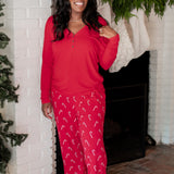 Smiling female model standing in front of a white brick fireplace wearing the Women's Lounge Pants in Candy Cane and Women's Henley Tee in Cardinal