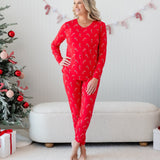 Female model standing with ankles crossed and one hand on hip wearing the Women's Jogger Pajama Set in Candy Cane between two decorated Chistmas trees and a cream ottoman
