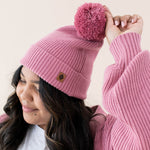 Focus is on the Pom on the hat and Kyte Baby's Leather Logo for the Chunky Knit Adult Pom Beanie in Apple Blossom