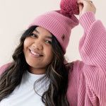 Smiling model wearing the Chunky Knit Adult Pom Beanie in Apple Blossom with one hand holding up the pom
