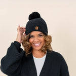 Model wearing  the Chunky Knit Adult Pom Beanie in Midnight with one hand on the brim of the beanie