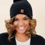 Smiling model wearing the Chunky Knit Adult Pom Beanie in Midnight with the Kyte Baby Leather Logo visible