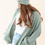 Model wearing the Chunky Knit Adult Pom Beanie in Thyme paired with a white shirt and Thyme Chunky Knit Oversized Women's Cardigan and jeans
