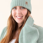 Smiling model wearing the Chunky Knit Adult Ribbed Beanie in Thyme with the Kyte Baby Leather logo visible