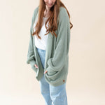 Model wearing the Chunky Knit Women's Oversized Cardigan in Thyme with a white shirt, light wash jeans and a Thyme Beanie