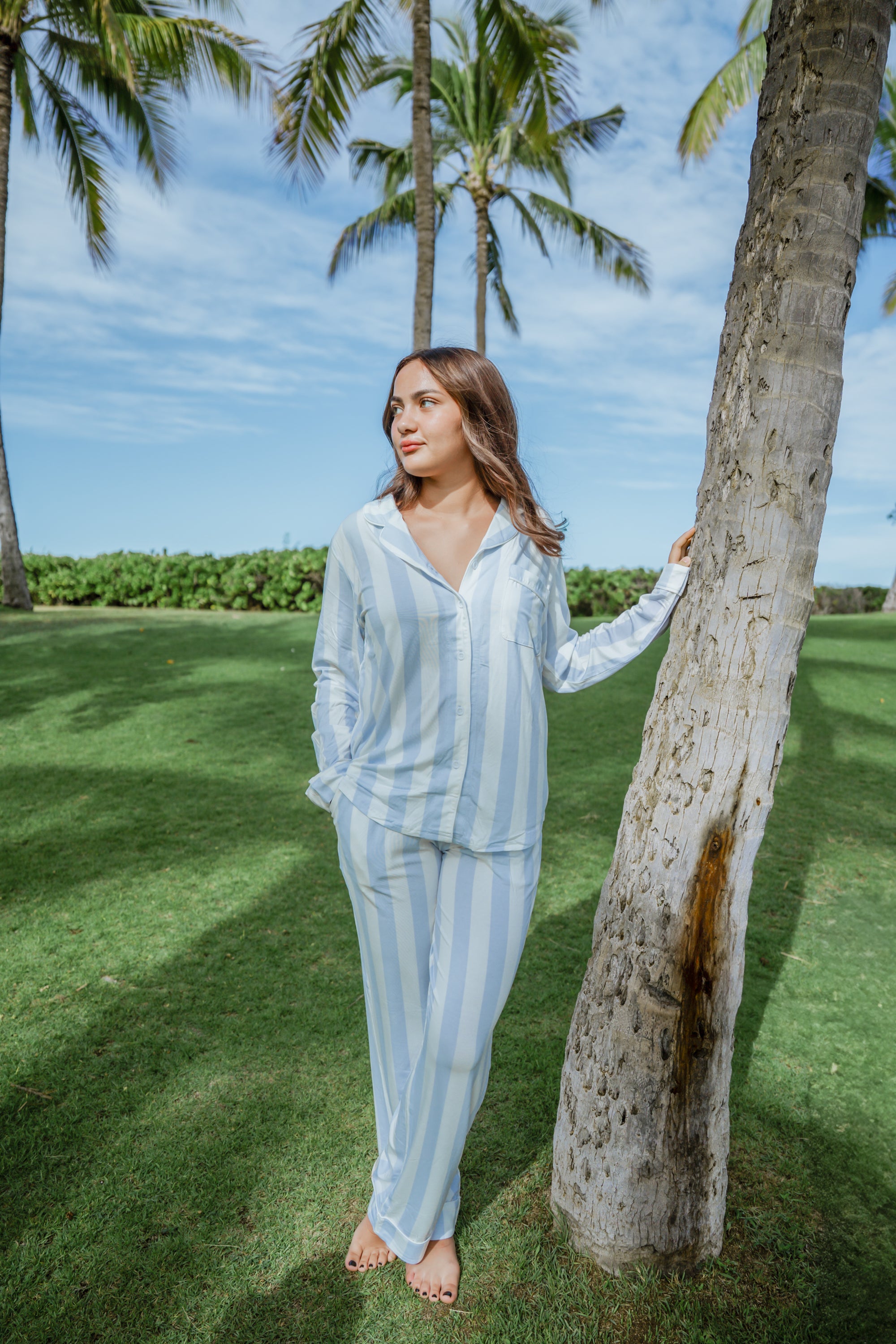 Woman in light blue striped pajama set standing next to a palm tree on a grassy area.