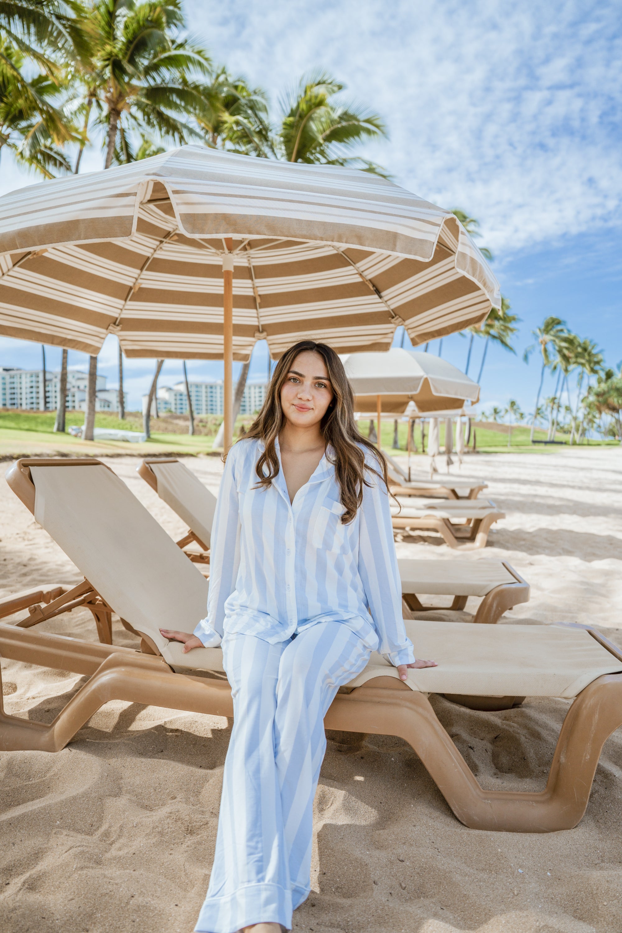 Woman in a light blue outfit sitting on a beach chair under a striped umbrella with palm trees and a clear sky in the background.