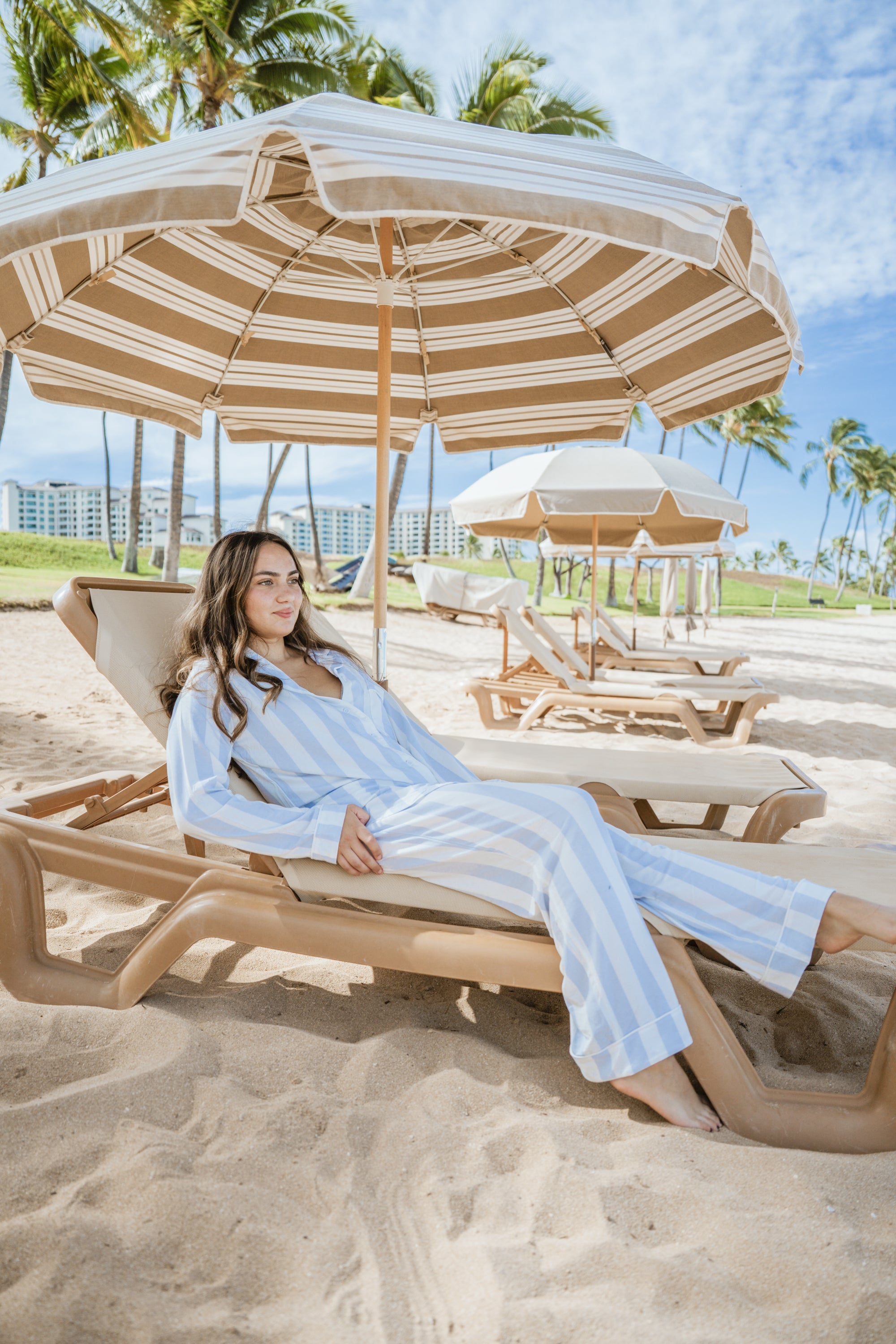 Woman relaxing on a beach chair under a large umbrella with palm trees and ocean in the background