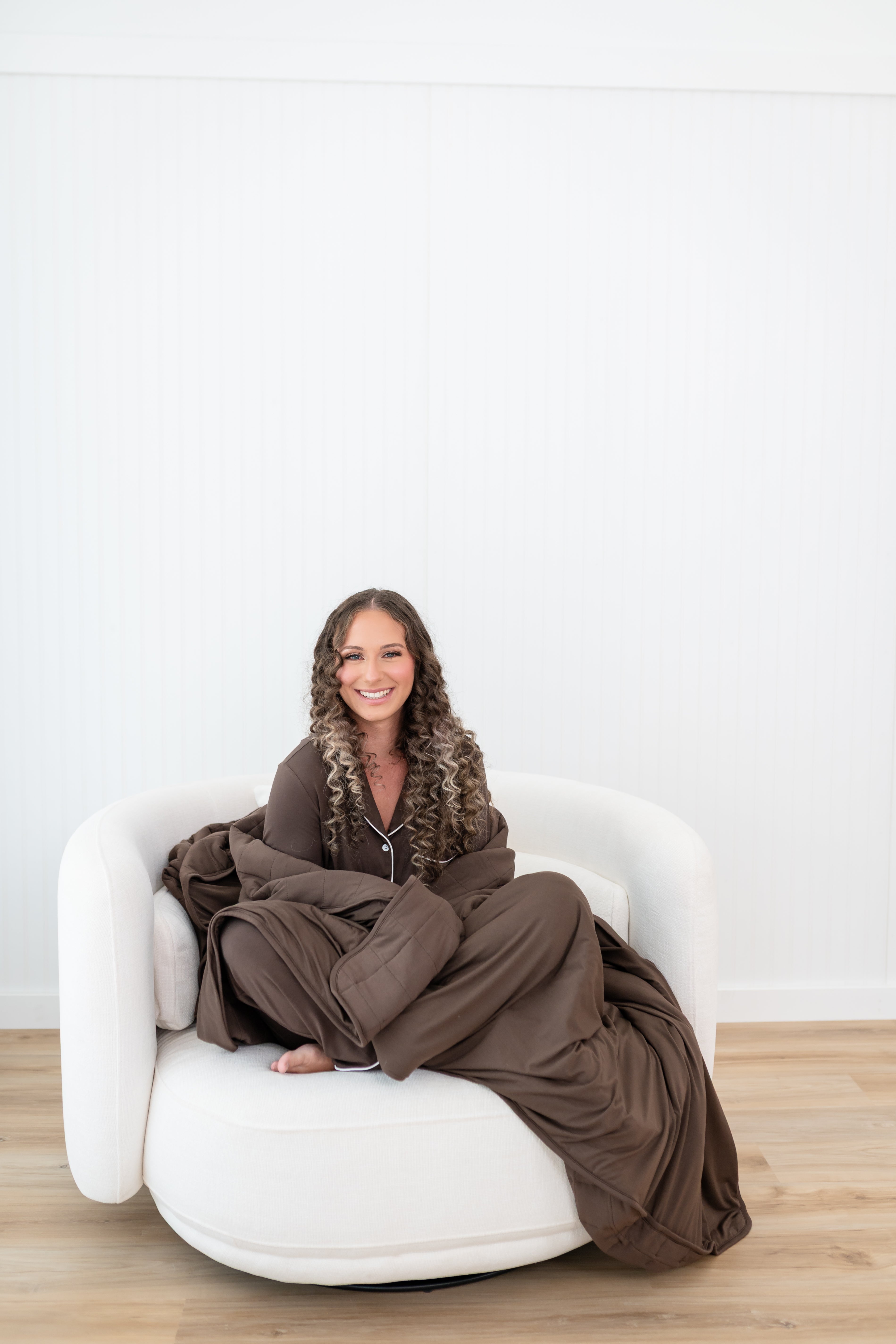 Smiling model curled up while wrapped in Model curled up on a white sofa in Kyte Quilt Blanket in Espresso 2.5 and matching pajama set