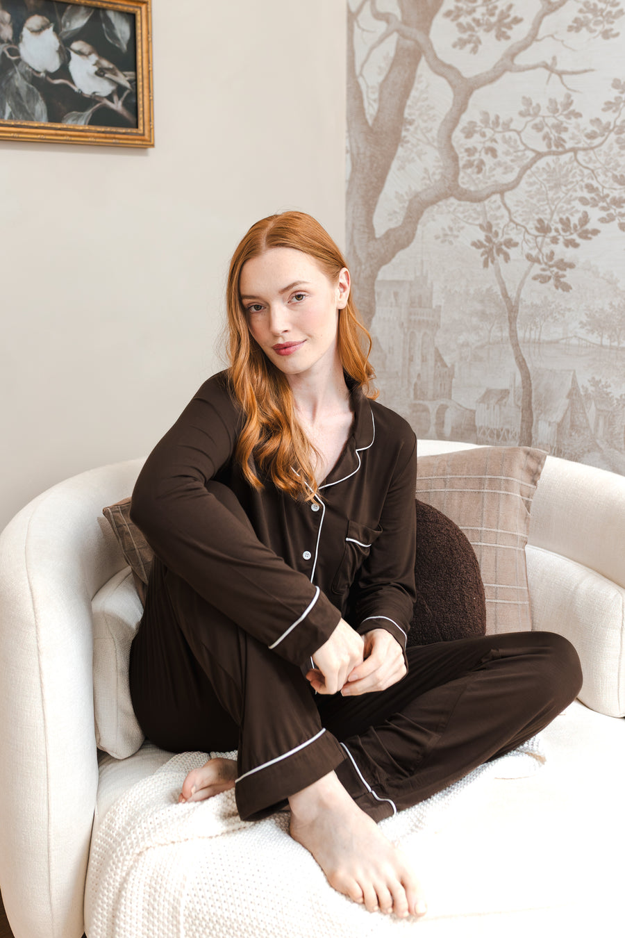 A woman with red hair sits cross-legged on a white armchair wearing a dark brown pajama set with white piping. The background features a beige wall with a framed bird painting and a gray tree-patterned wallpaper.