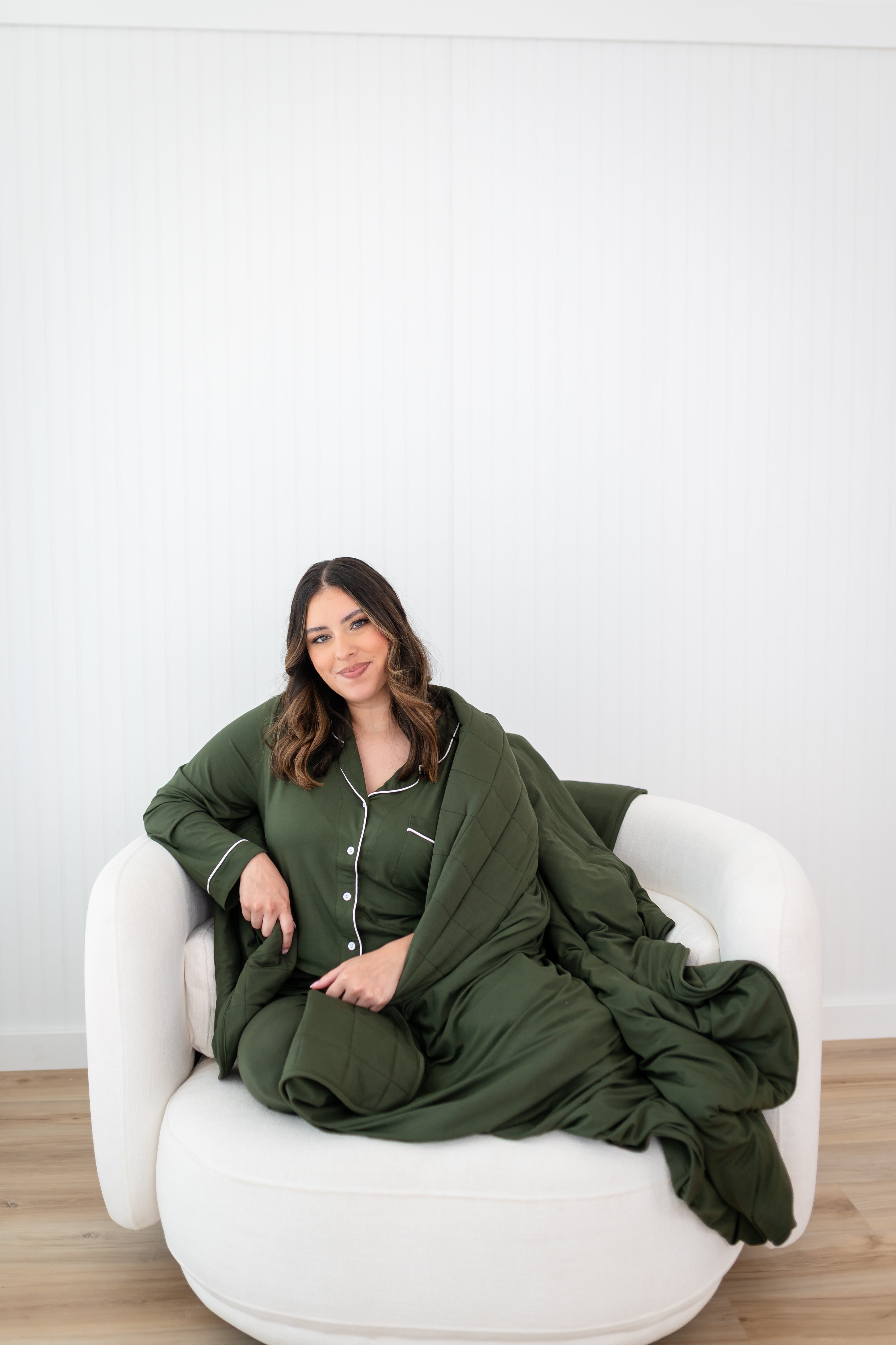 Model sitting on a white couch sofa wrapped in a the bamboo Quilt Blanket in Fir 2.5 and wearing the matching Long Sleeve Pajama Set