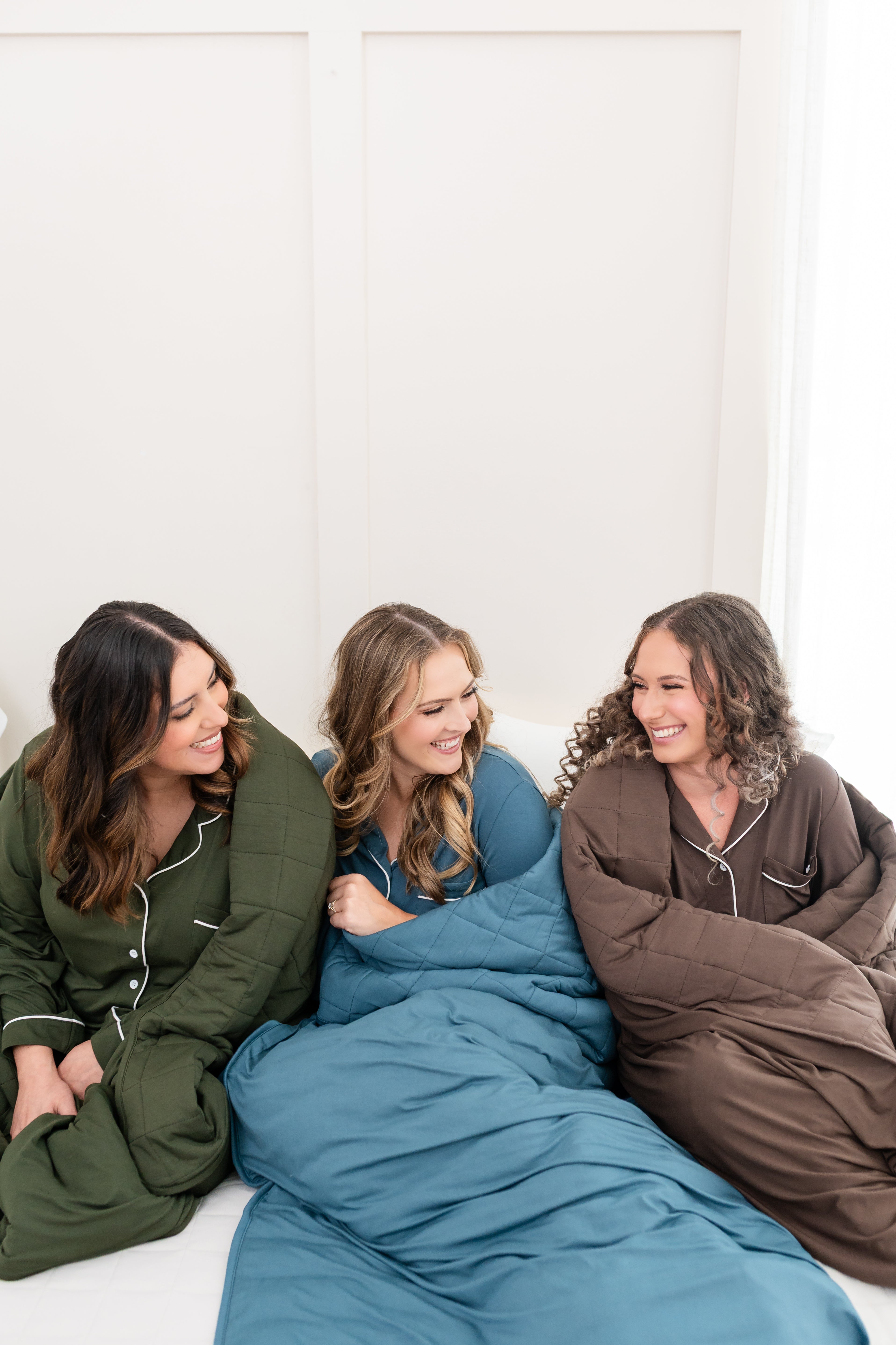 Models snuggled on a bed wrapped in Quilted Blankets in Fir, Atlantic and Espresso