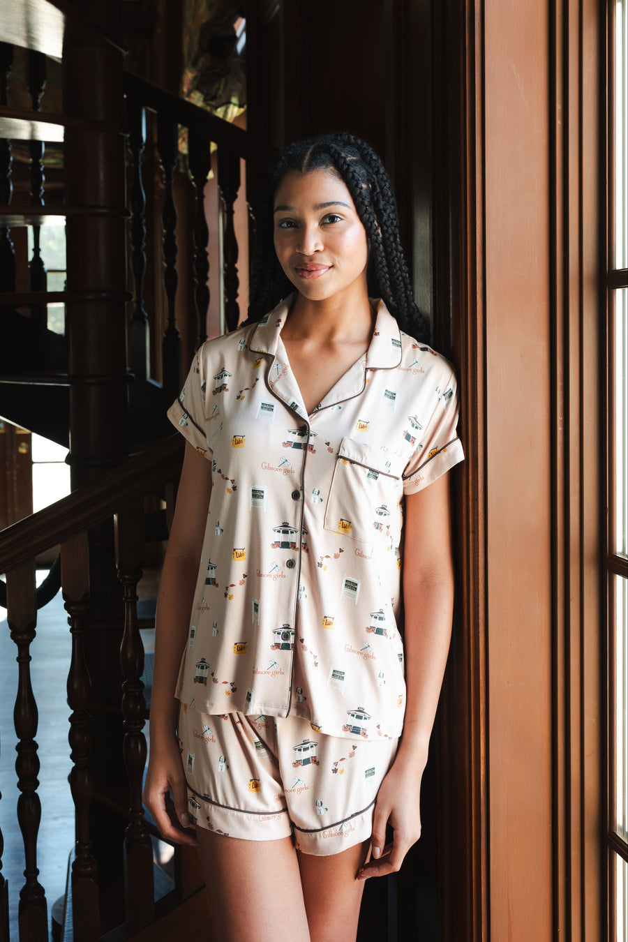 A woman wearing a short-sleeve pajama shirt and matching shorts with a small illustrated Gilmore Girls print, standing indoors near a wooden staircase and window.