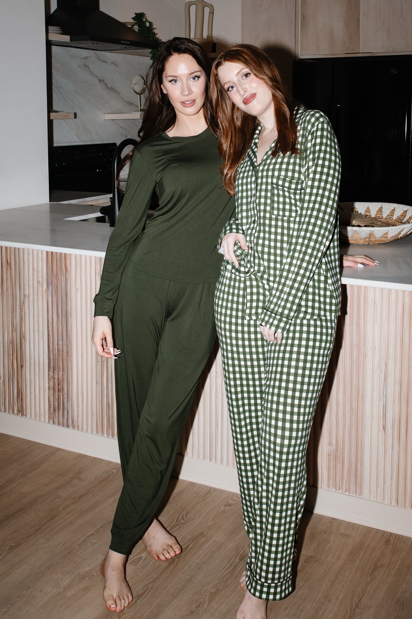 Models standing next to a counter while wearing the Women's Long Sleeve Pajama Set in Gingham Fir and Jogger Set in Fir