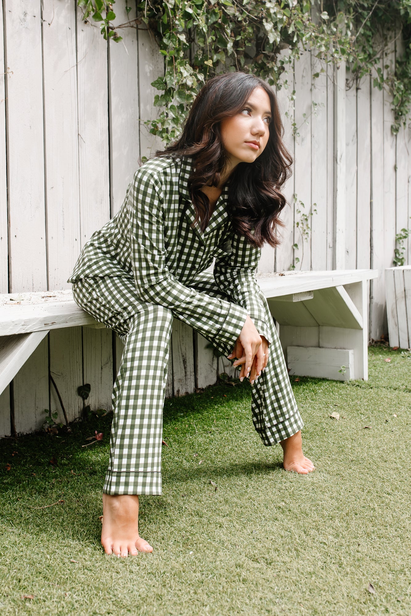Woman in green checkered pajamas sitting on grass next to a wooden bench.