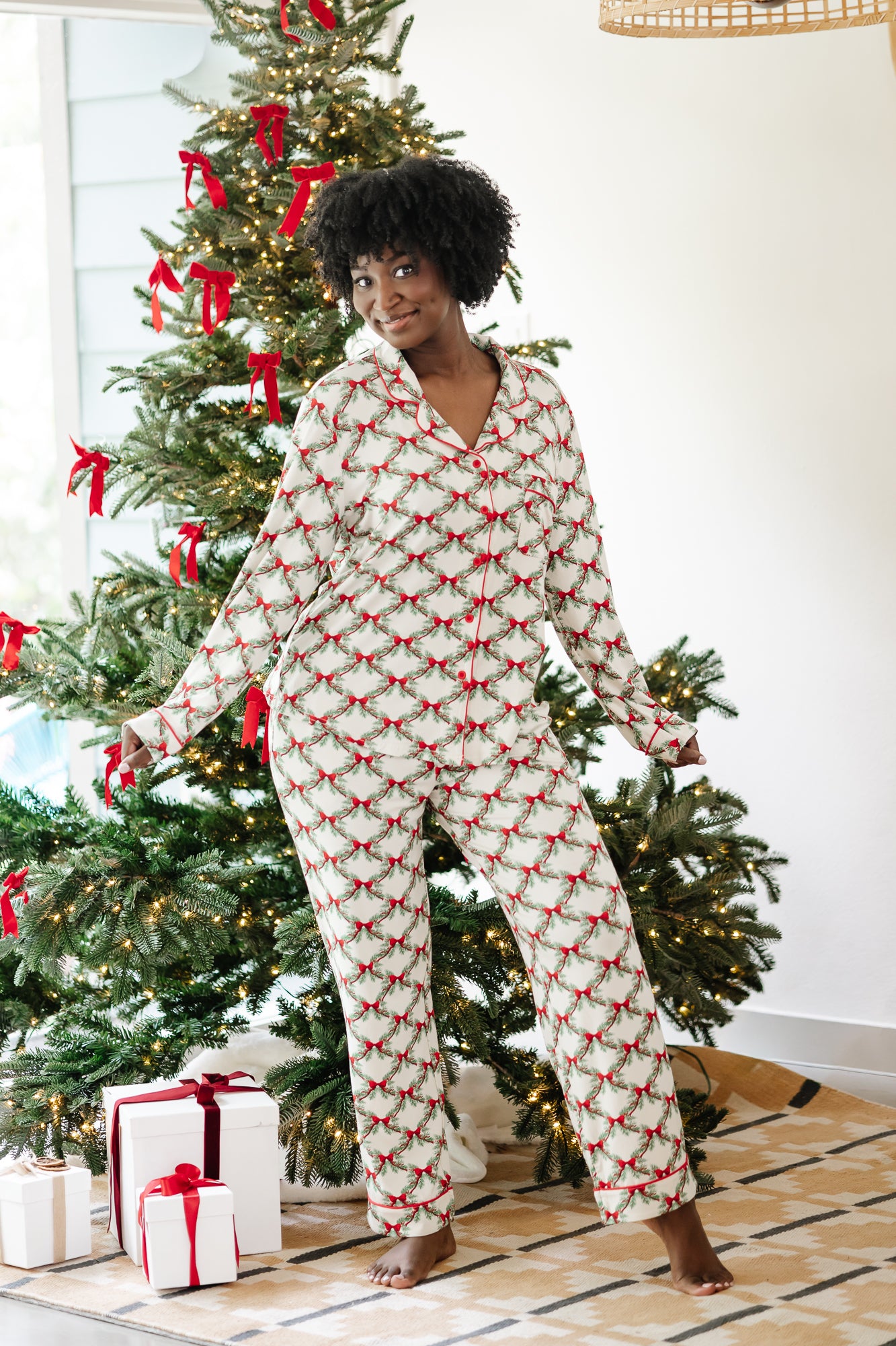 Smiling model standing in front of a Christmas tree wearing the Long Sleeve Women's PJ Set in Holiday Bow