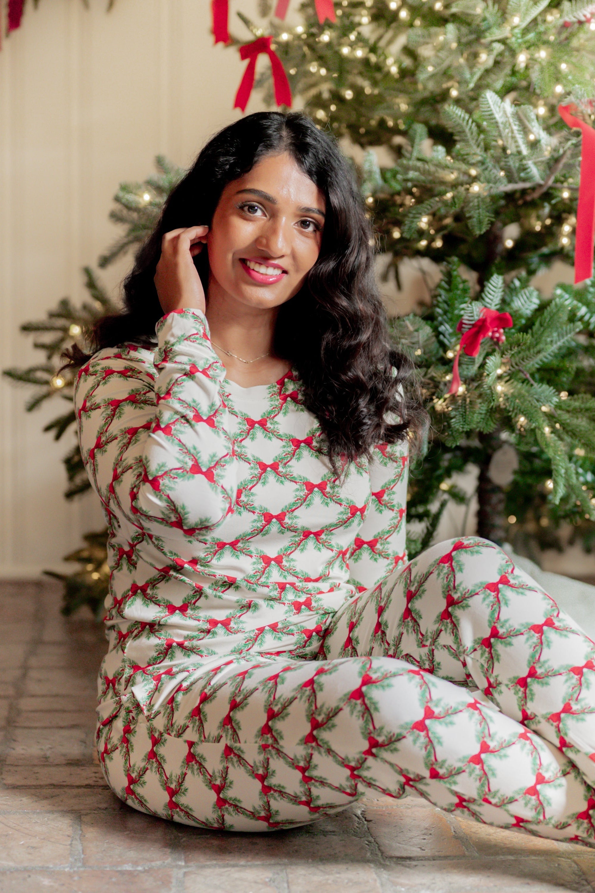 Smiling female model sitting on the floor wearing the Women's Jogger Pajama Set in Holiday Bow in front of a decorated Christmas Tree