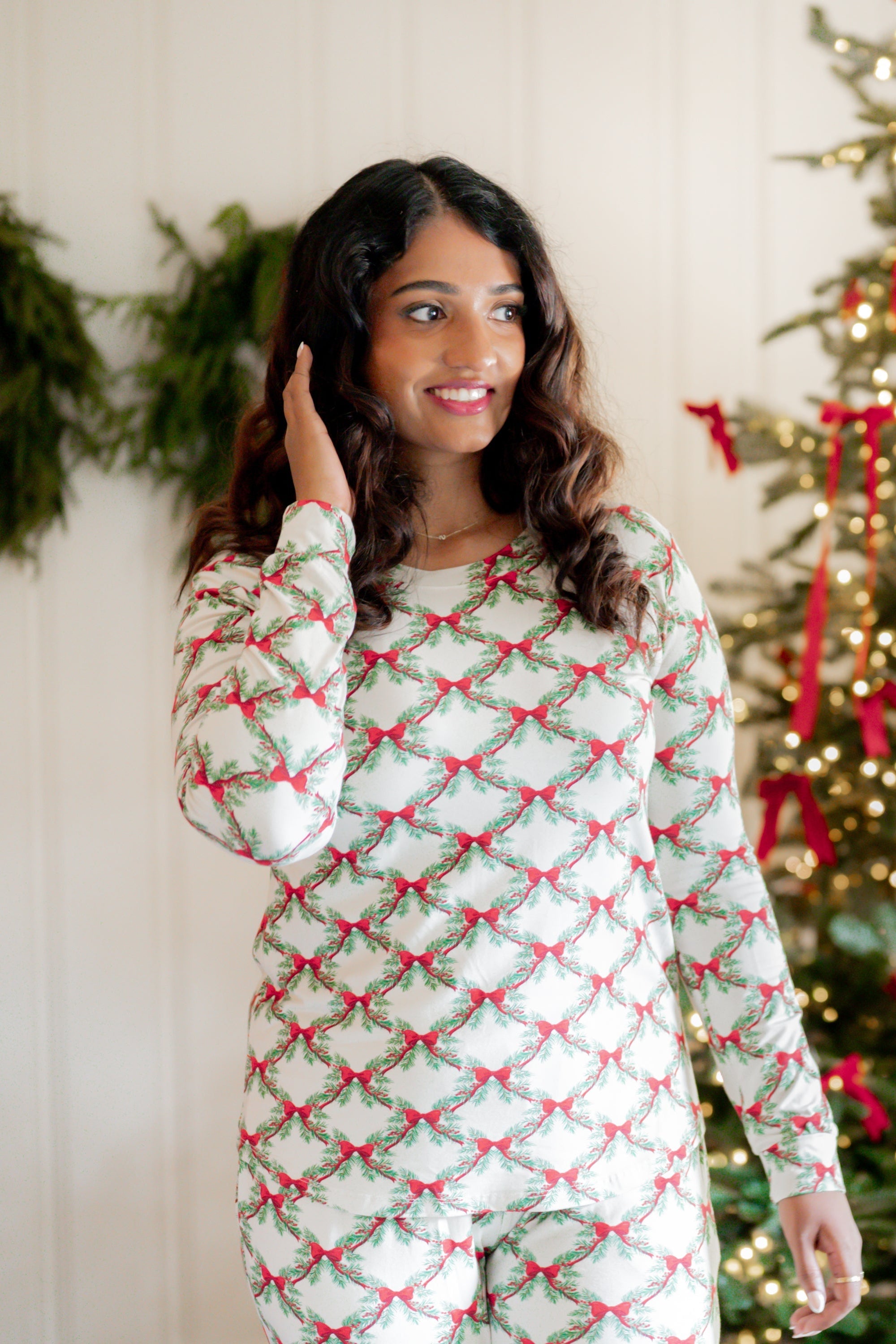 Close up of the top of the Women's Jogger Pajama Set in Holiday Bow shown on a model standing in front of a white paneled wall and decorated Christmas tree