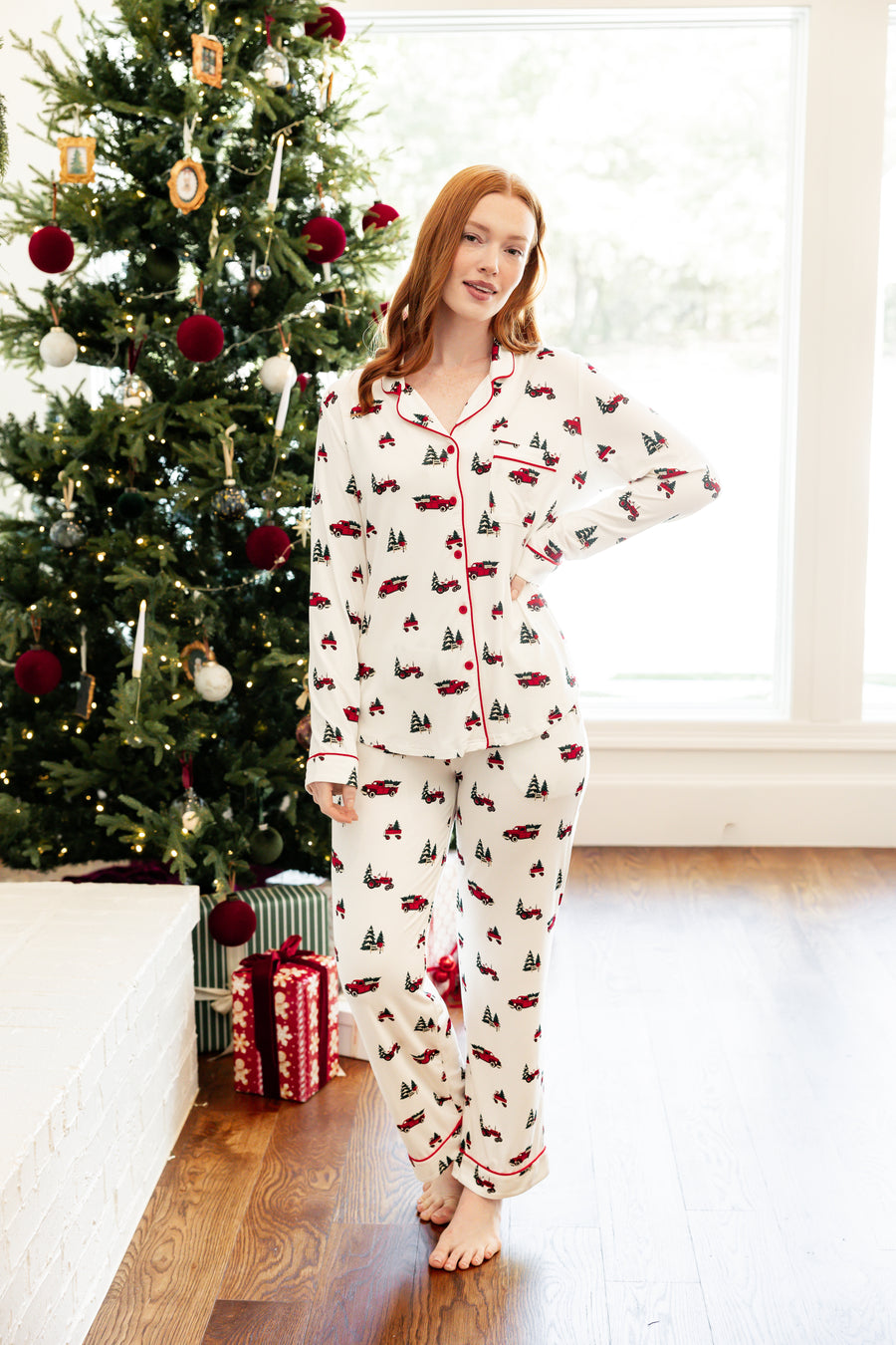 A woman wearing a matching holiday-themed pajama set stands barefoot in a living room next to a decorated Christmas tree. The pajamas are off-white with a repeating pattern of red trucks, green trees, and red accents.