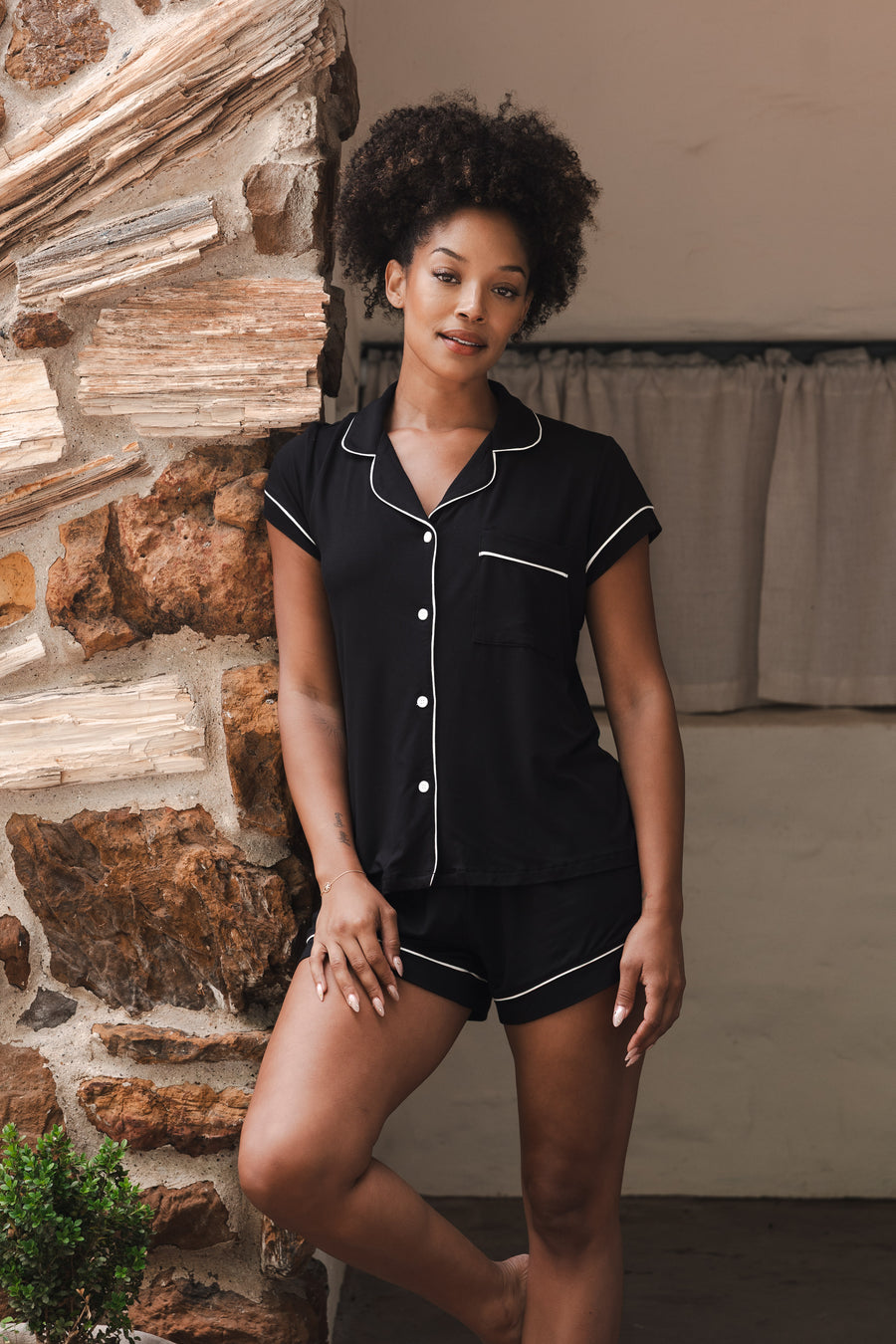 Woman wearing a black pajama set with white trim against a stone wall.