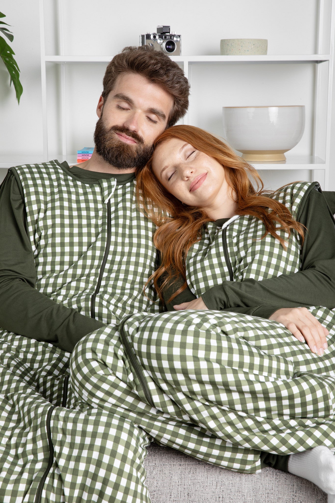 Two people wearing green and white checkered sleep bag walkers sitting on a couch.