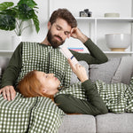 Two people in matching green checkered sleep bag walkers lying on a couch, with one person holding a book.