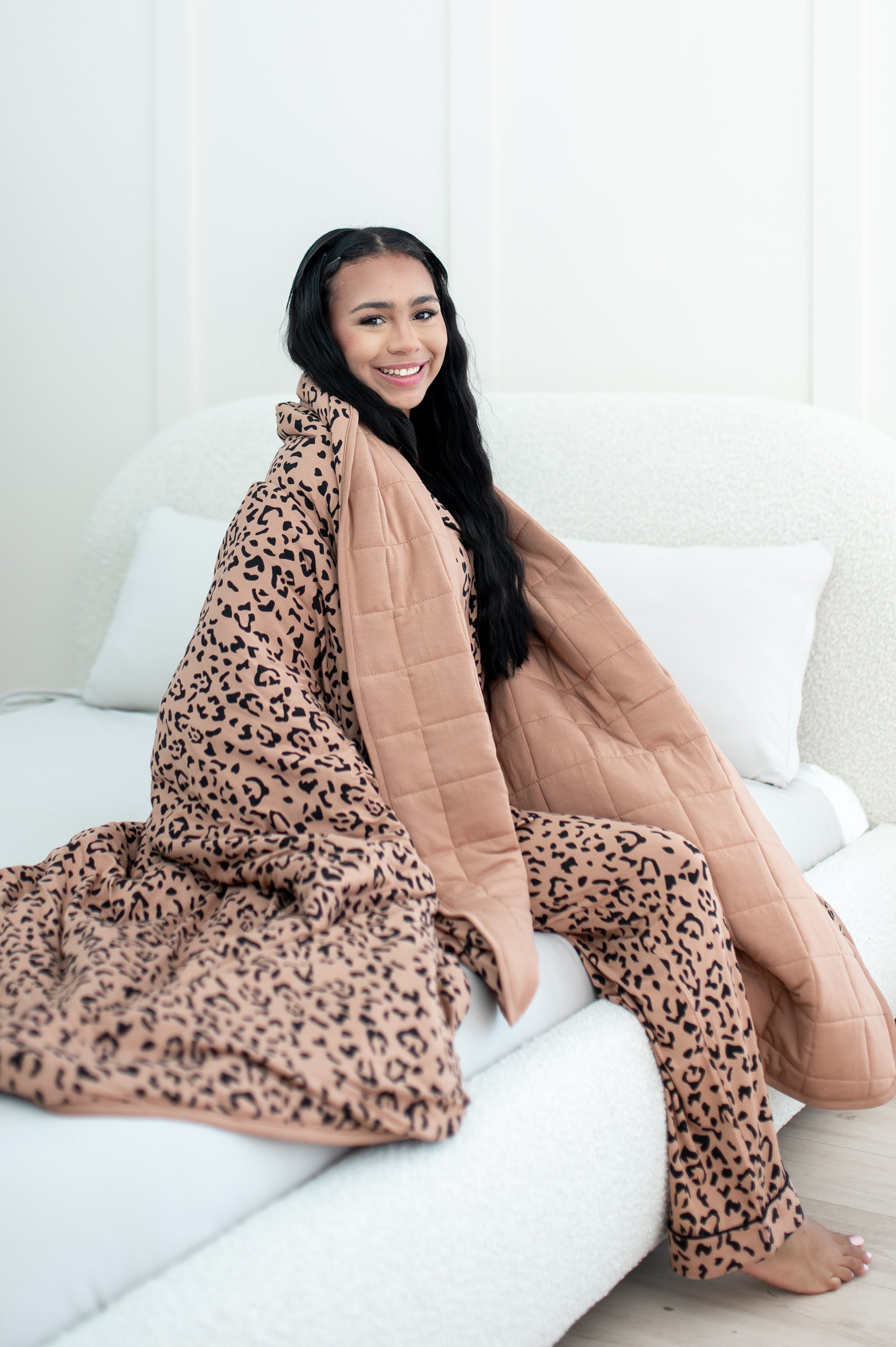 Smiling model sitting on a white bed wrapped in Kyte Quilt Blanket in Latte Leopard 2.5