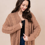 Model smiling downwards wearing the Chunky Cable Knit Cardigan in Latte