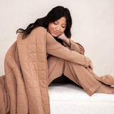 Model sitting wrapped up in the Bamboo Quilt Blanket in Latte 2.5 while wearing matching pajamas