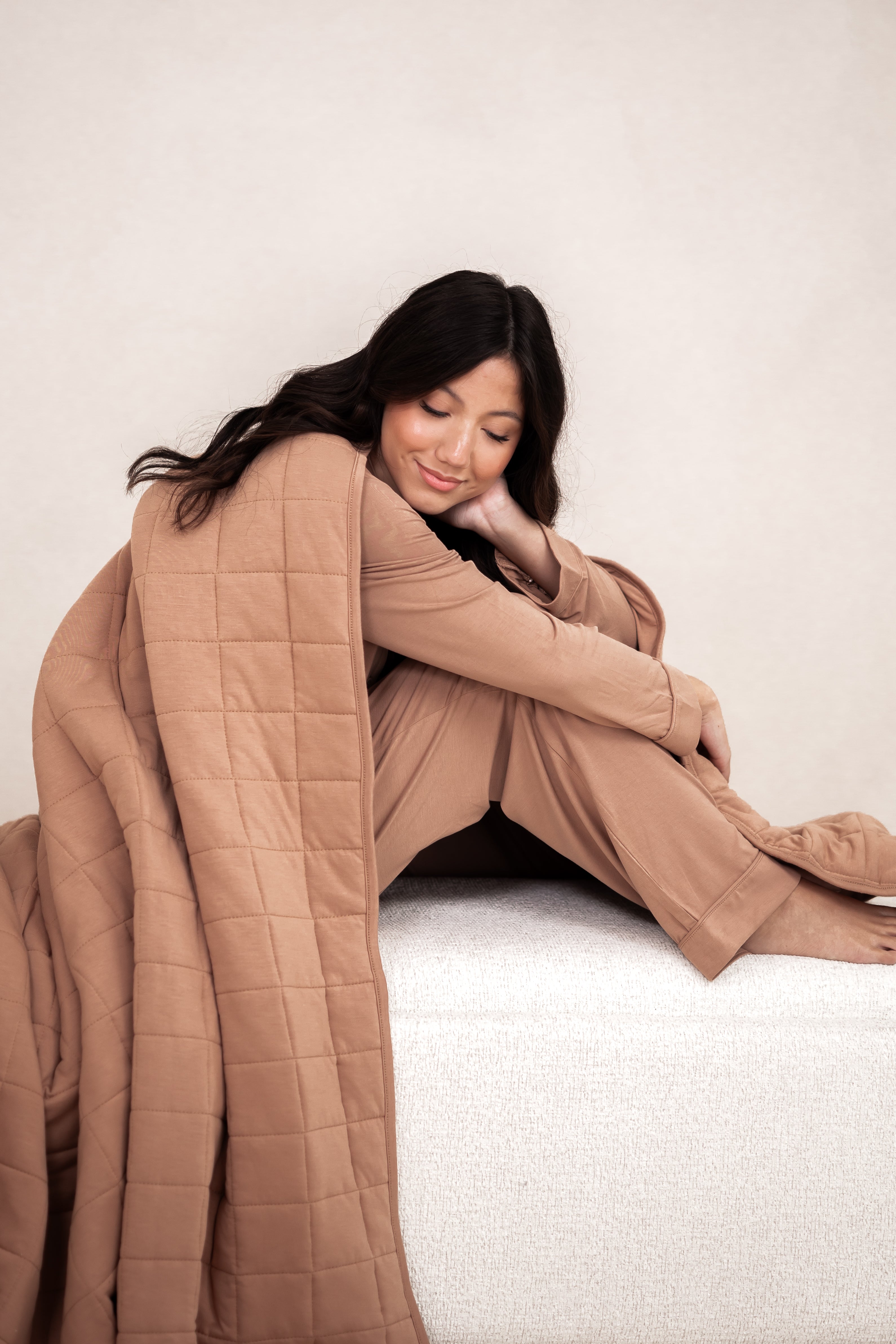 Model sitting wrapped up in the Bamboo Quilt Blanket in Latte 2.5 while wearing matching pajamas