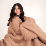 Model sitting wrapped in the Kyte Bamboo Quilt Blanket in Latte 2.5 and looking away