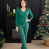 Smiling female standing between two Christmas trees wearing the Women's Lounge Pants in Merry and Bright paired with an Emerald Women's Henley tee