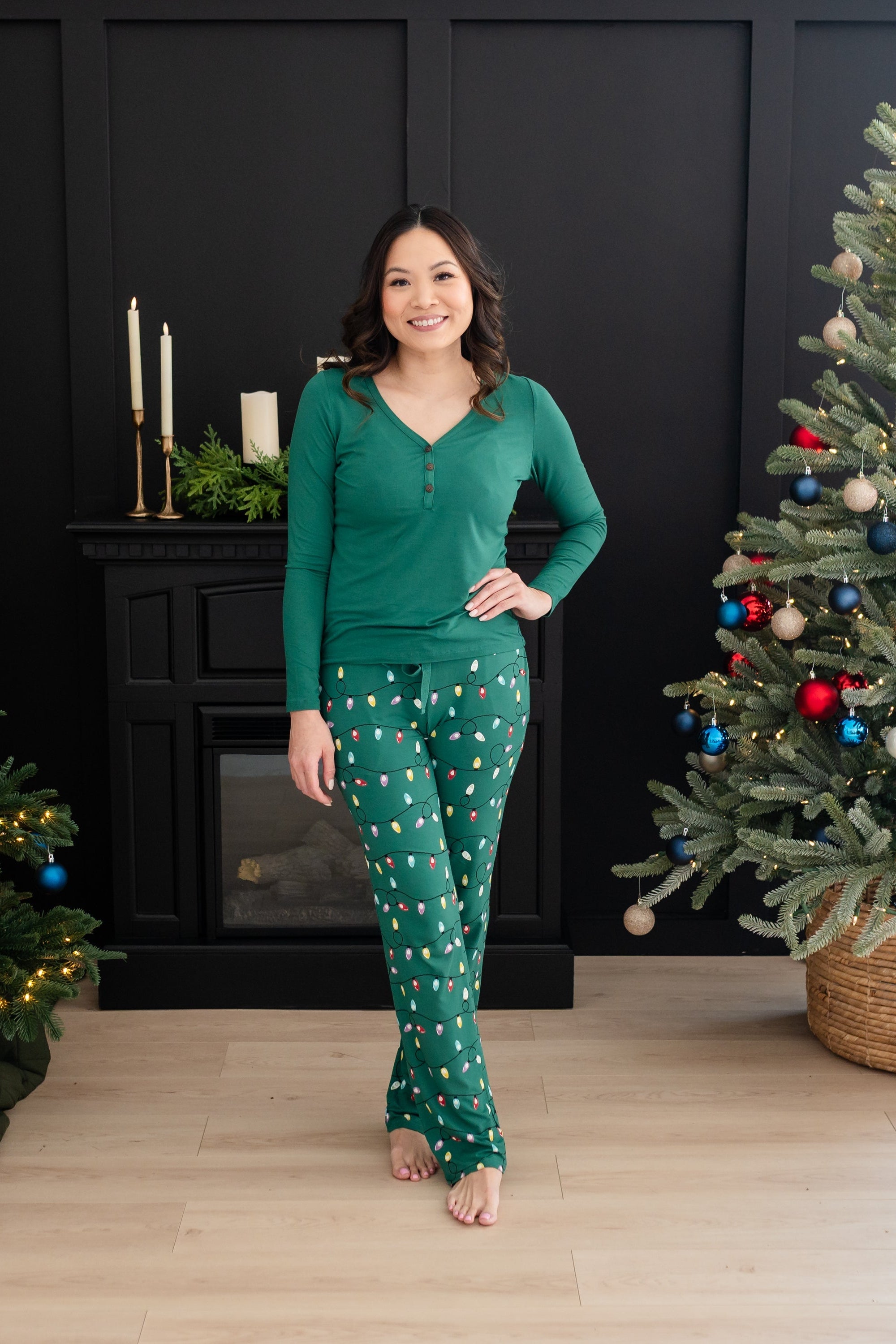 Smiling female standing between two Christmas trees wearing the Women's Lounge Pants in Merry and Bright paired with an Emerald Women's Henley tee