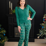 Smiling model standing between two Christmas trees with hand on her hip wearing the Women's Lounge Pants in Merry and Bright and Emerald Long sleeve Henley tee