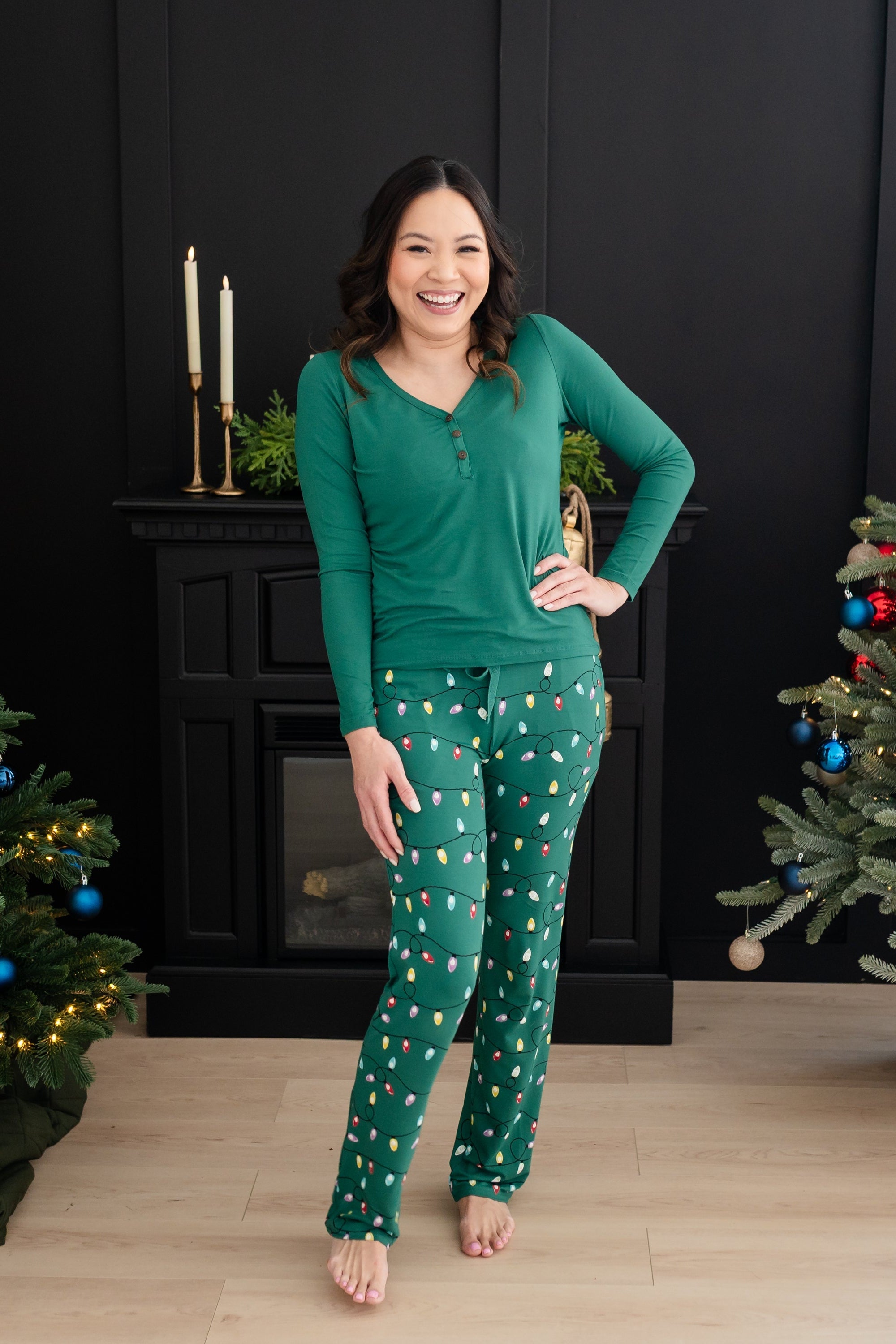 Smiling model standing between two Christmas trees with hand on her hip wearing the Women's Lounge Pants in Merry and Bright and Emerald Long sleeve Henley tee